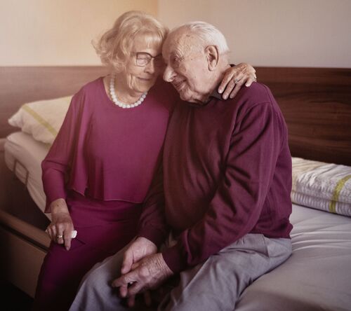 Älteres Paar sitzt vertraut am Pflegebett und zeigt tiefe Zuneigung