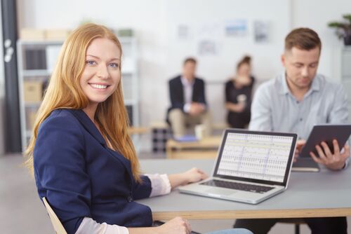 Frau in einem großen Büro sitzend an einem Tisch, auf dem ein Laptop steht, am selben Tisch und im Hintergrund weitere Personen bei der Arbeit